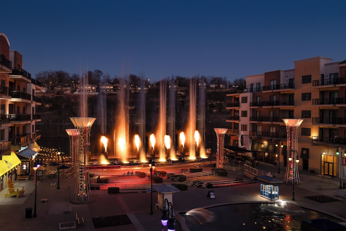 A nighttime view of a public square with a fountain, surrounded by modern buildings and street lamps.