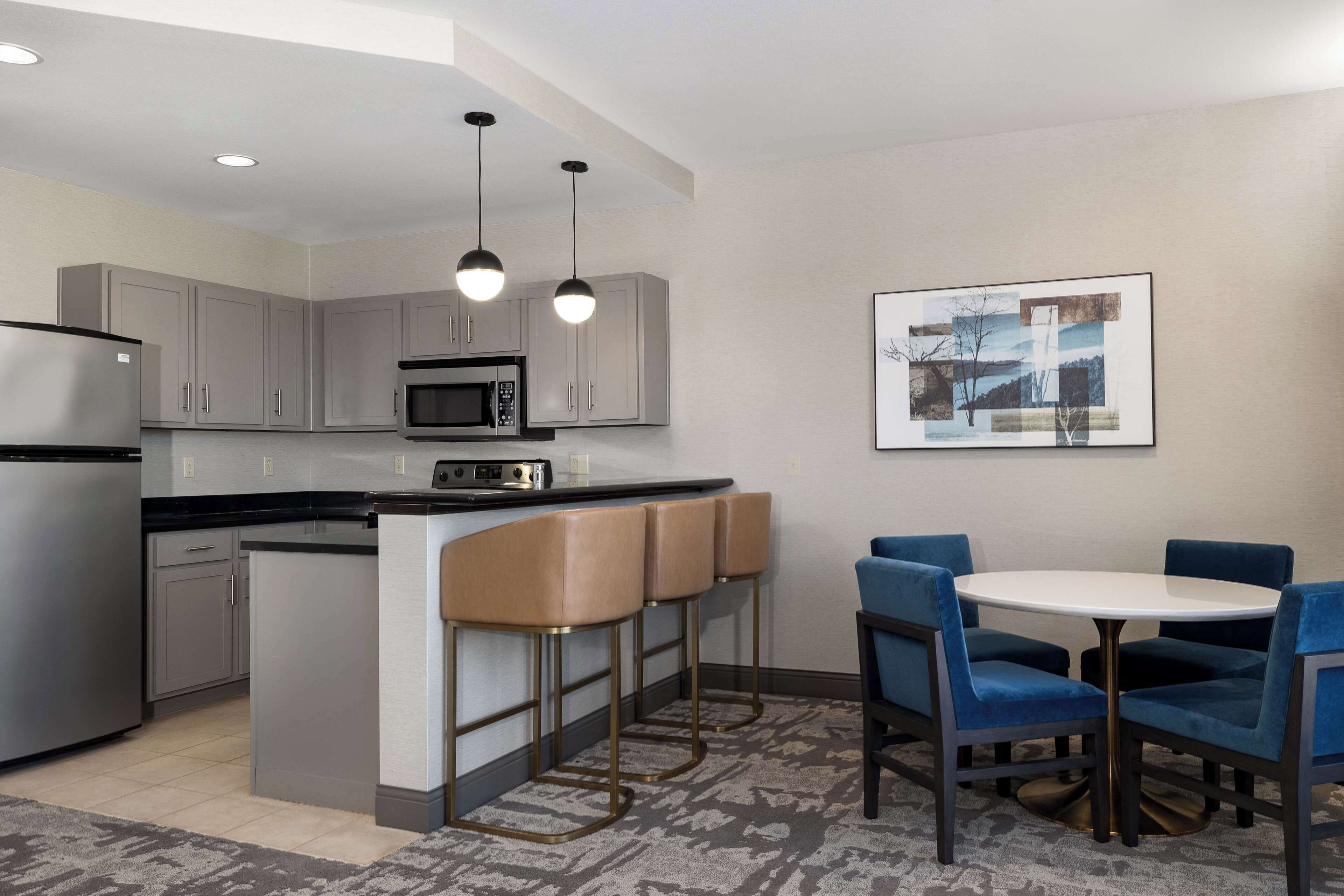 A modern kitchen with grey cabinets, a stainless steel refrigerator, a microwave, and a bar area with stools. There is a round table with blue chairs and a framed picture on the wall.