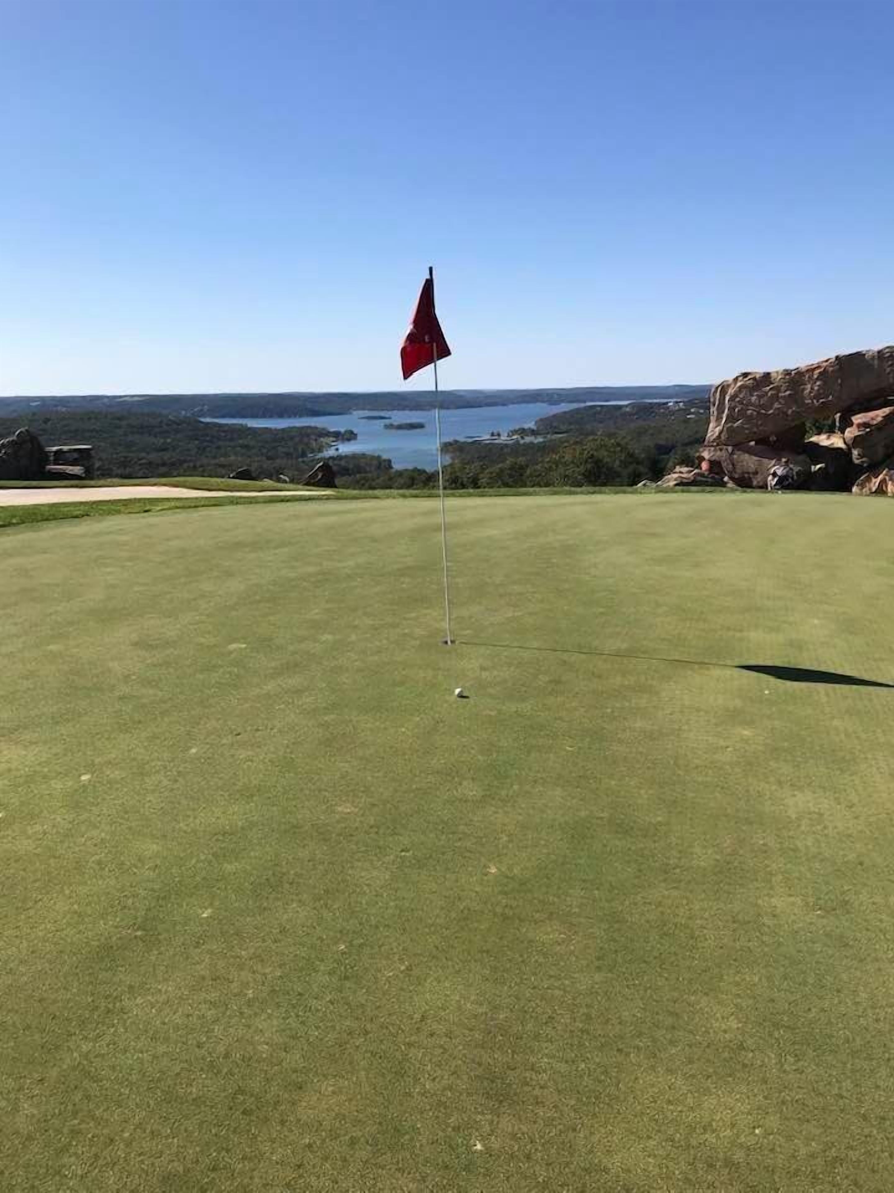 A golf course with a red flag on the green, surrounded by rocky terrain and a view of a lake.