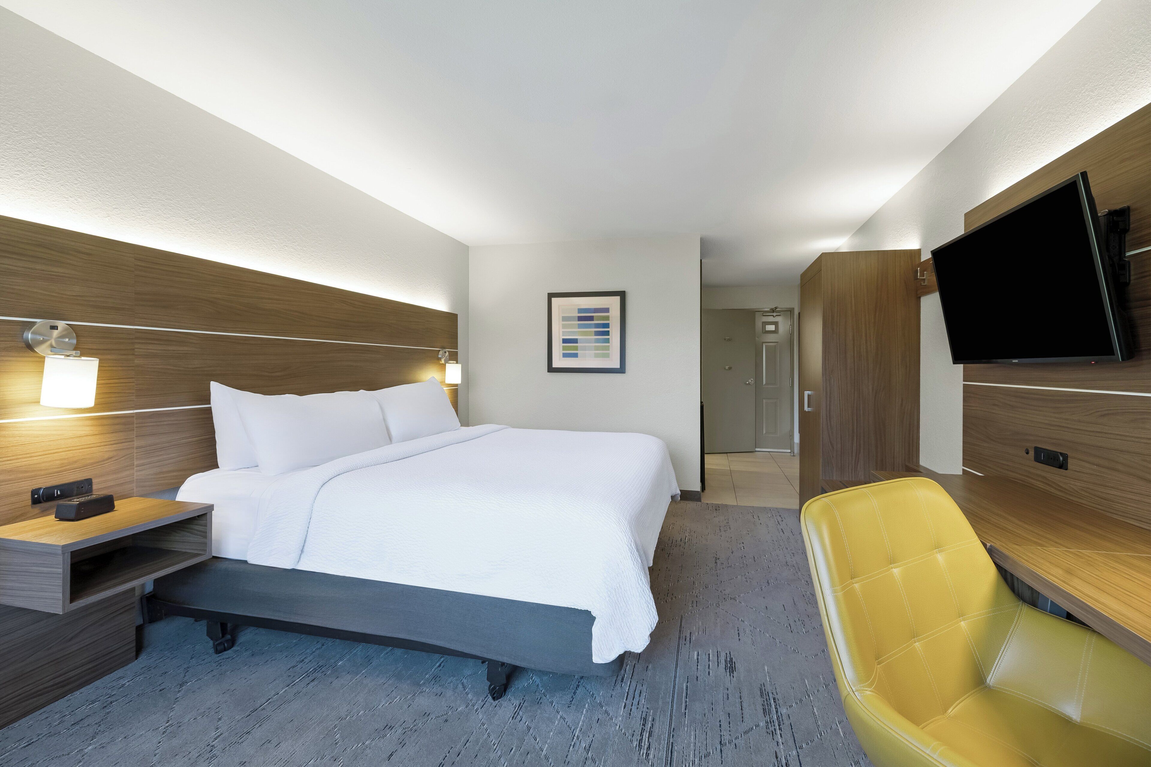 Una habitación de hotel con cama, mesita de noche, lámpara, televisor y una silla amarilla.