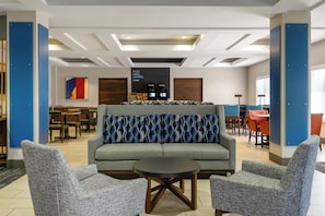 A modern lounge area with a sofa, two armchairs, a coffee station, and a dining area with tables and chairs.