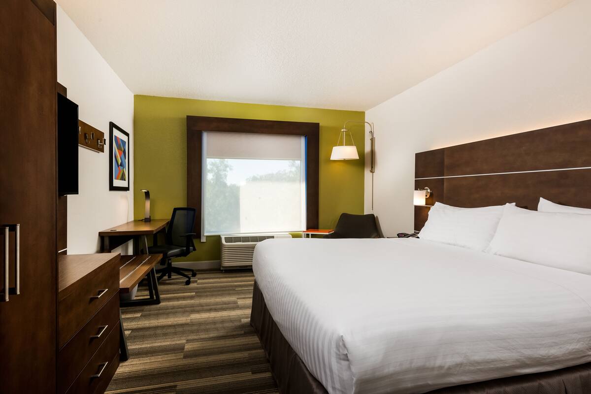 A hotel room with a large bed, a desk with a chair, a wardrobe, a window with blinds, and a wall-mounted lamp.