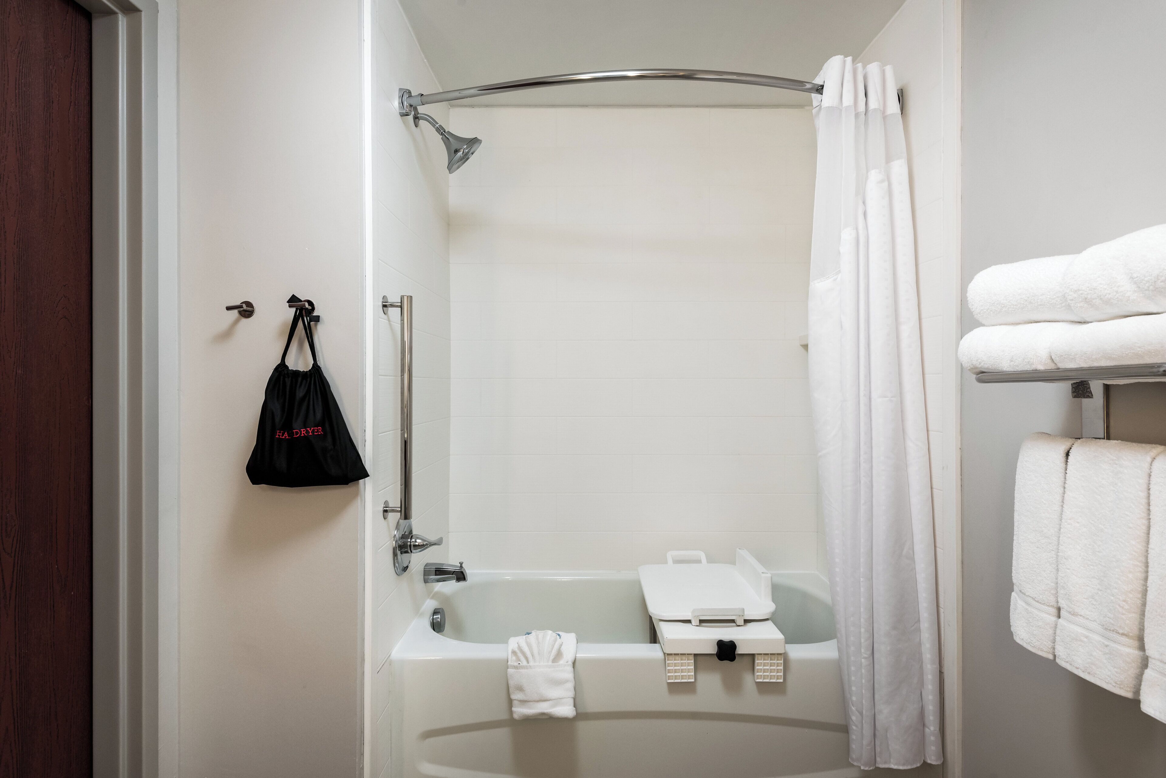 A bathroom with a bathtub, a shower curtain, a towel rack with towels, and a black bag hanging on the wall.