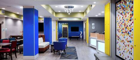 Vestíbulo de hotel con decoración azul y amarilla, zonas de estar, una mesa de recepción y una pantalla de televisión.