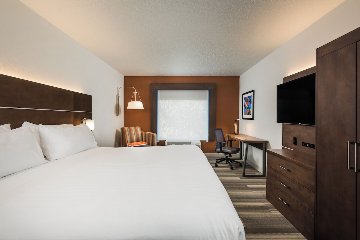 A hotel room with a large bed, a desk with a chair, a television, and a window with blinds.