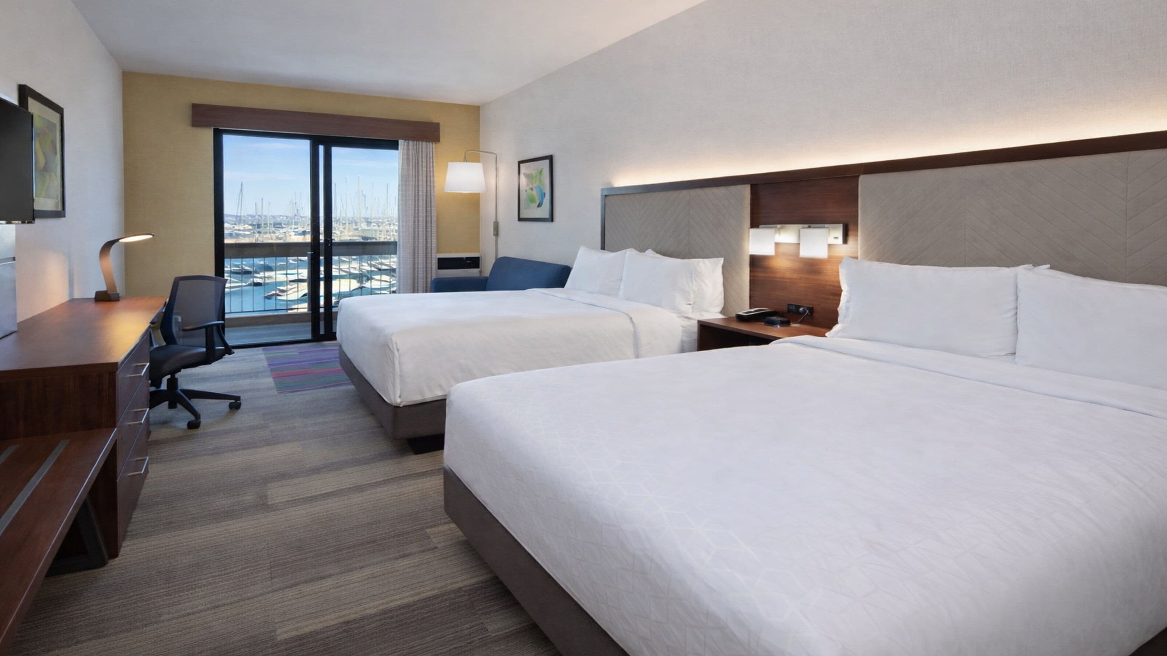 A hotel room with two beds, a desk, a chair, and a view of the marina.