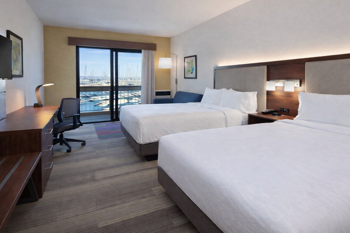 Een hotelkamer met twee bedden, een bureau, een stoel en uitzicht op de jachthaven.