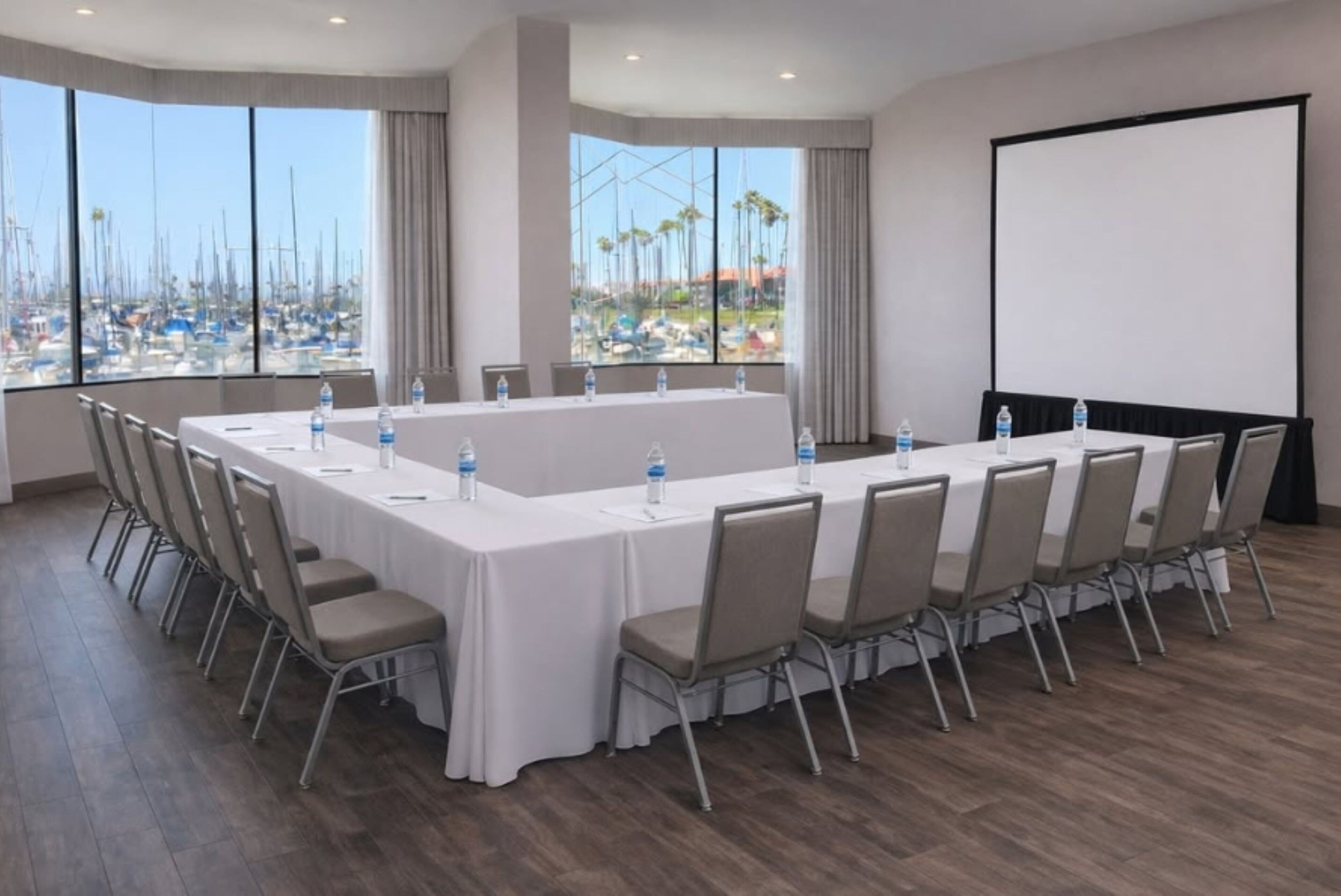 A conference room with a long table, chairs, a projector screen, and large windows with a marina view.