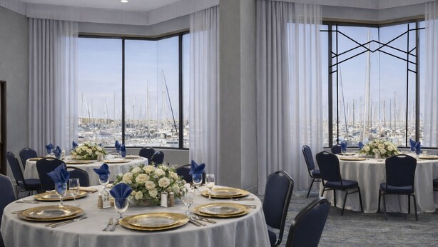 Comedor con mesas redondas dispuestas para una comida formal, ventanas amplias con vista al puerto deportivo y una lámpara con diseño geométrico.
