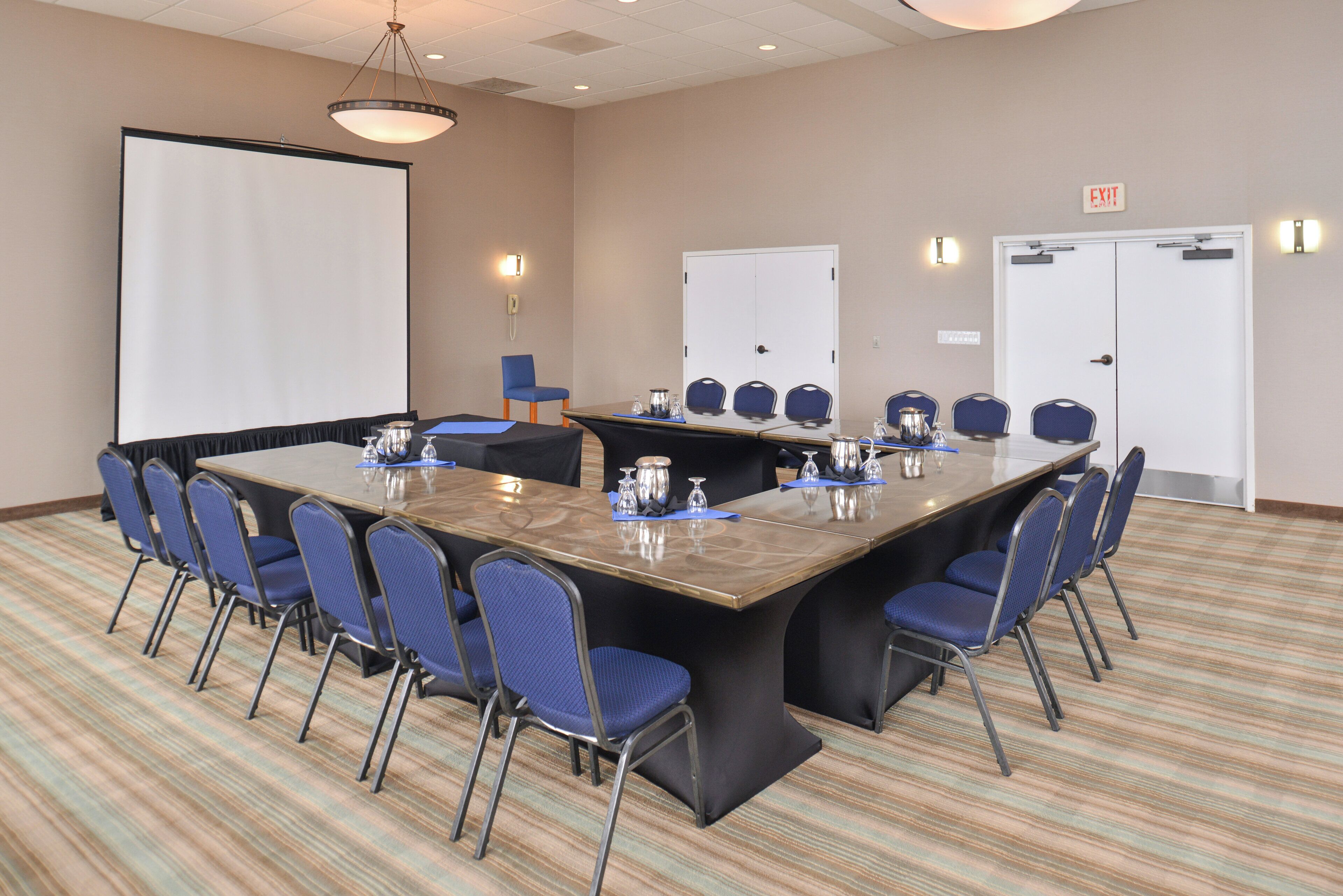 A conference room with a large table, blue chairs, a projector screen, and a door with an exit sign.