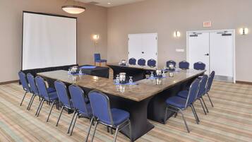 A conference room with a large table, blue chairs, a projector screen, and a door with an exit sign.