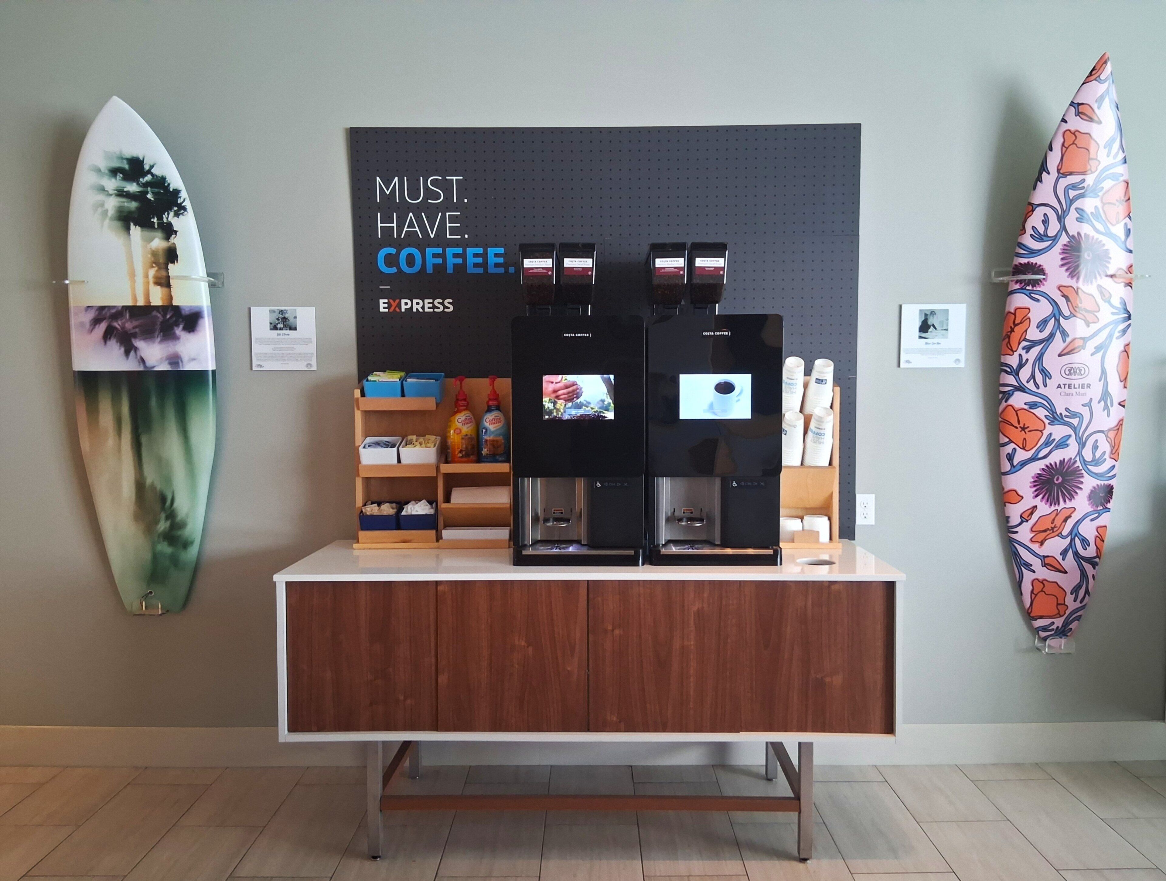 A coffee station with two espresso machines, a shelf with coffee pods, and a surfboard on the wall.