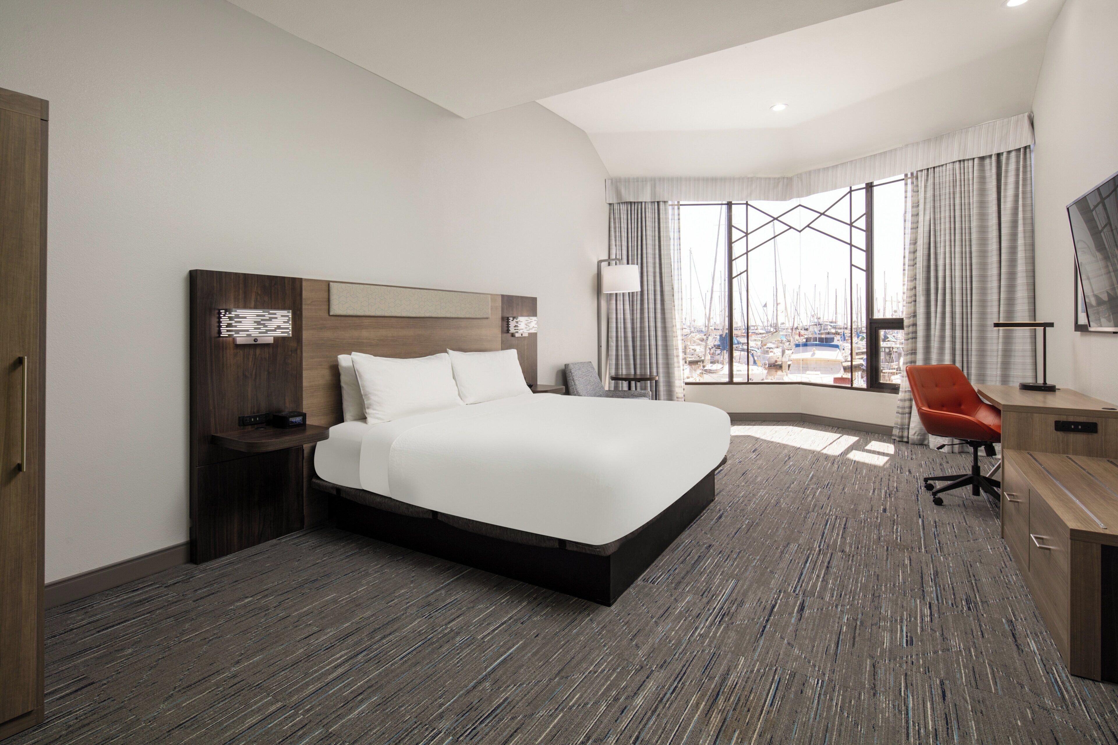 A modern hotel room with a large bed, a desk, and a view of boats through the window.