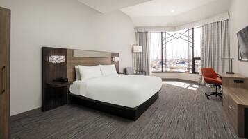 A modern hotel room with a large bed, a desk, and a view of boats through the window.