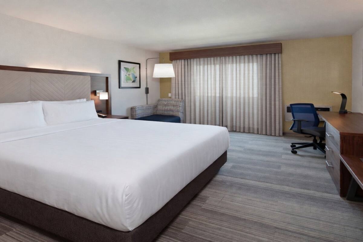 A hotel room with a large bed, a desk, a chair, a lamp, and a window with curtains.
