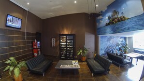 A lobby with a reception desk, black leather sofas, a coffee machine, a TV screen, and a mural of a beach scene. - Hotel Plaza Rosa (Medellin)