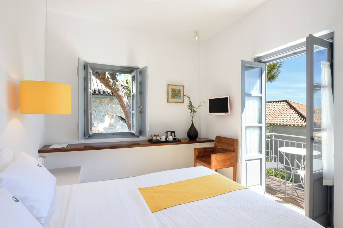 A hotel room with a bed, a desk with a lamp, a window with a view, a TV, and a balcony with outdoor furniture.