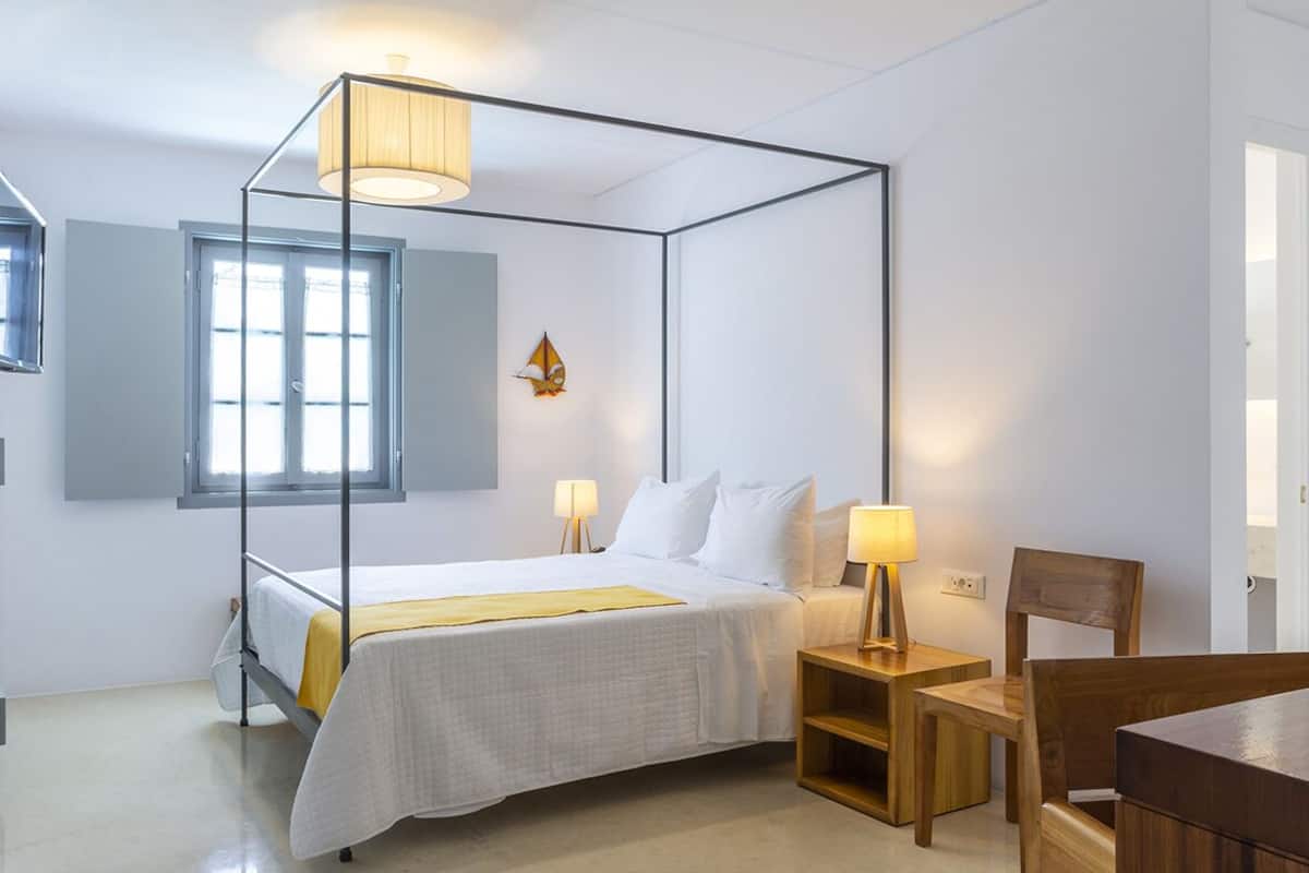 A four-poster bed with a canopy, a bedside table with a lamp, a wooden chair, and a window with curtains.