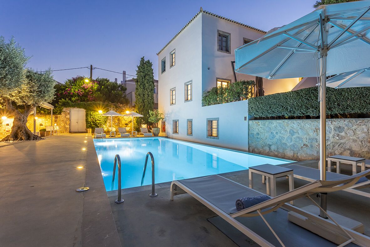 Piscina con sillones de descanso y sombrillas, rodeada por un edificio con muro de piedra y un árbol.