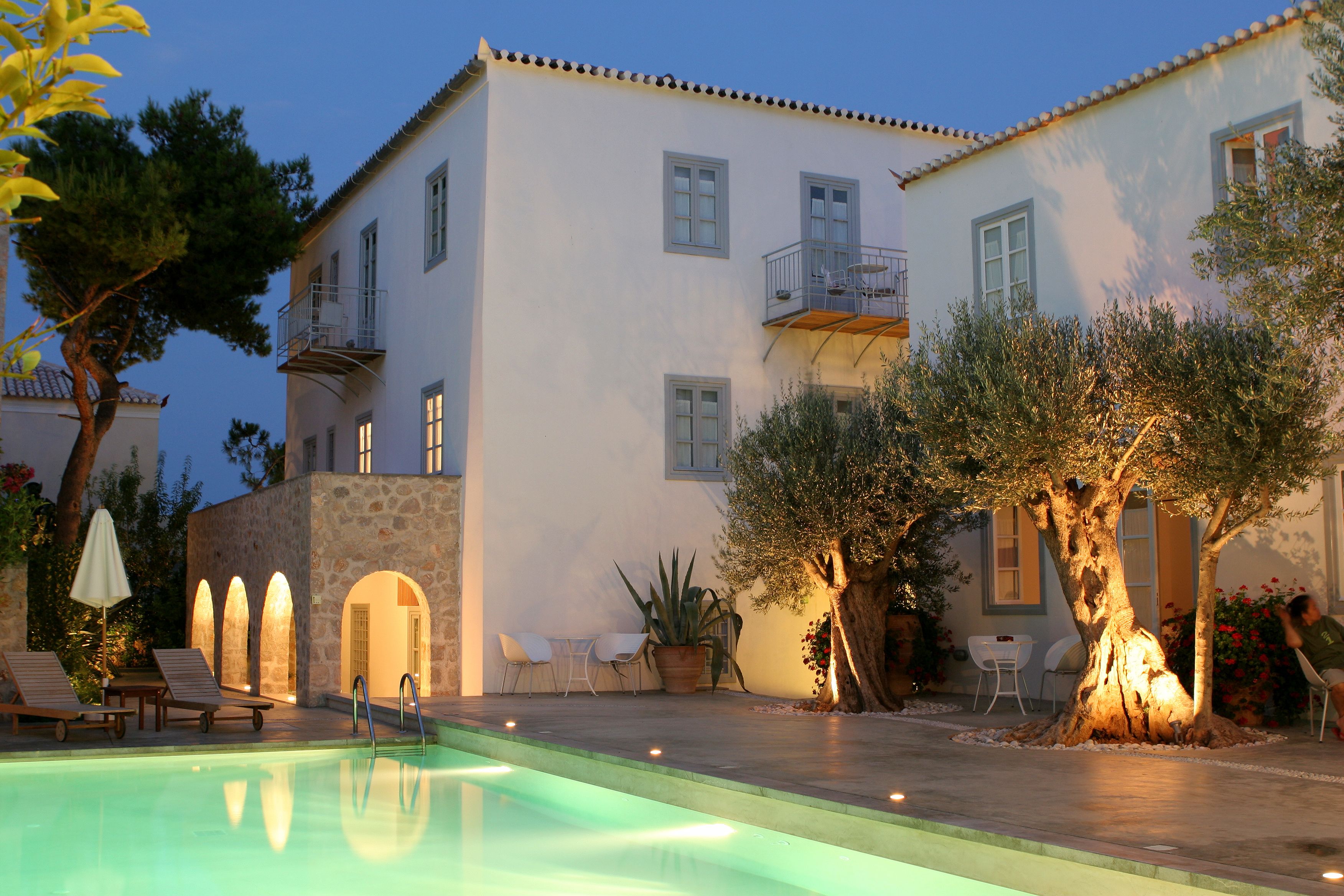 Un área junto a la piscina con sillones de descanso, un muro de piedra y un edificio con balcones y ventanas.
