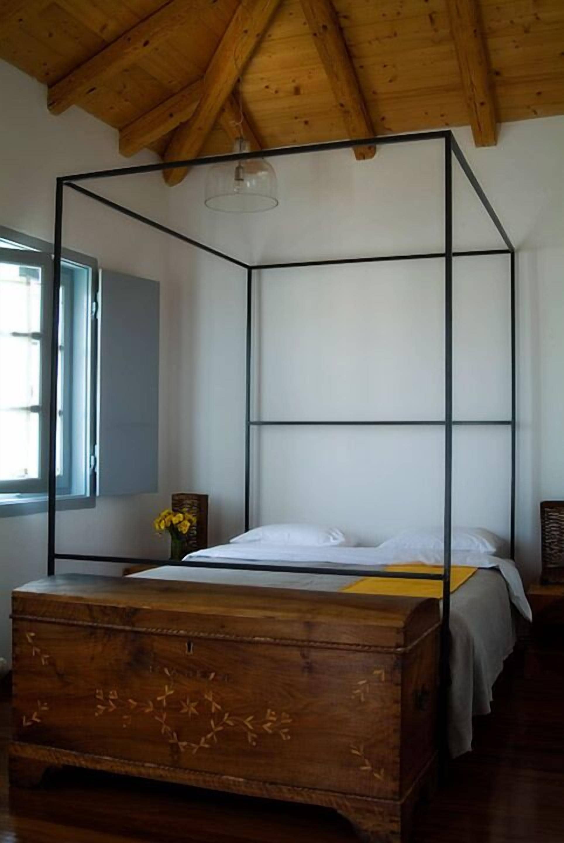 A wooden four-poster bed with a white mattress and pillows, a wooden nightstand with a vase of flowers, and a window with a view of the outside.