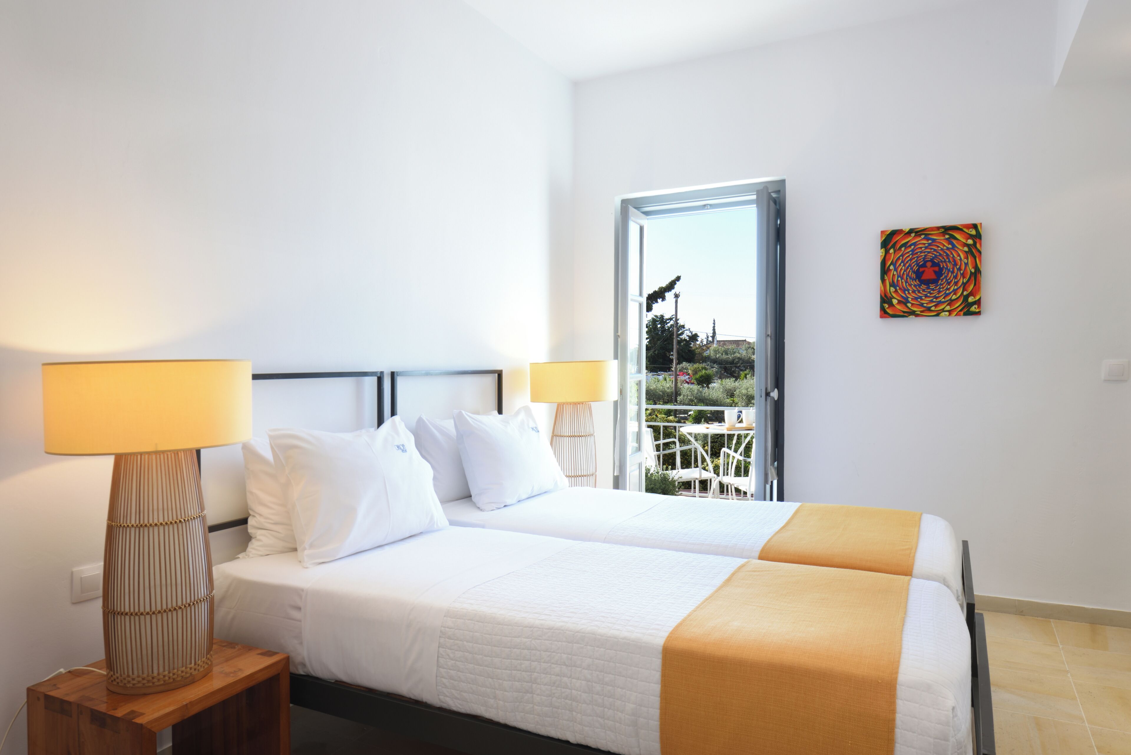A hotel room with two beds, a window with a view of greenery, a table with chairs outside, and a colorful artwork on the wall.