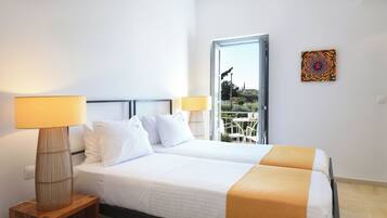 A hotel room with two beds, a window with a view of greenery, a table with chairs outside, and a colorful artwork on the wall.