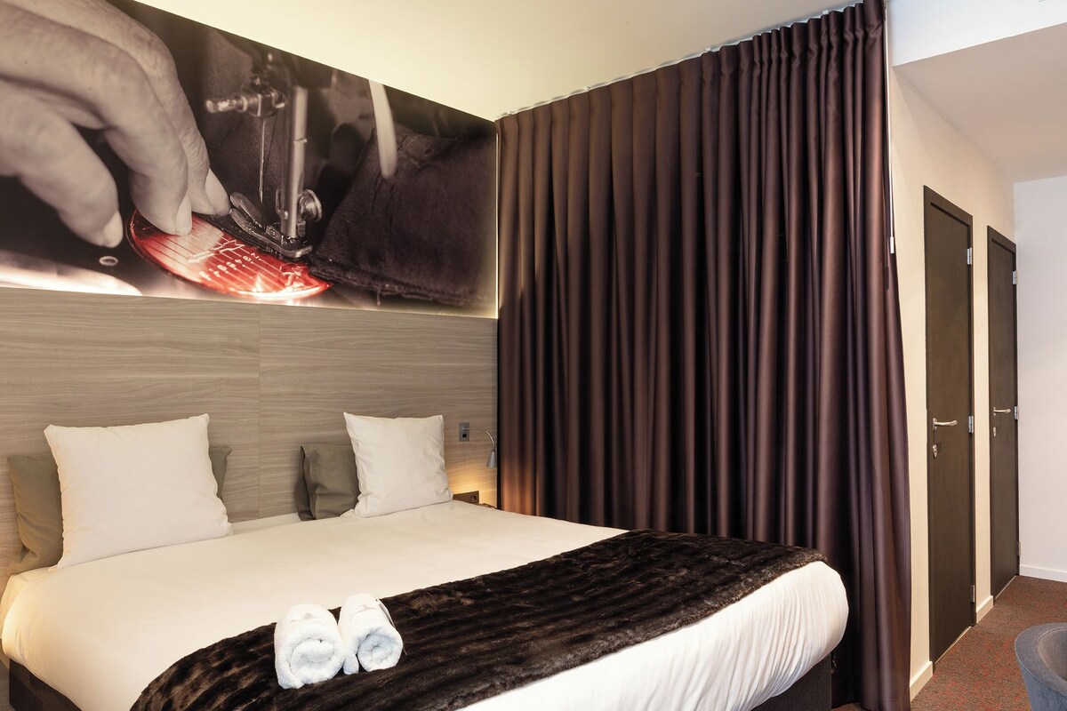 Habitación de hotel con una cama grande, un cabecero con un cartel, cortinas oscuras y una puerta a la derecha.