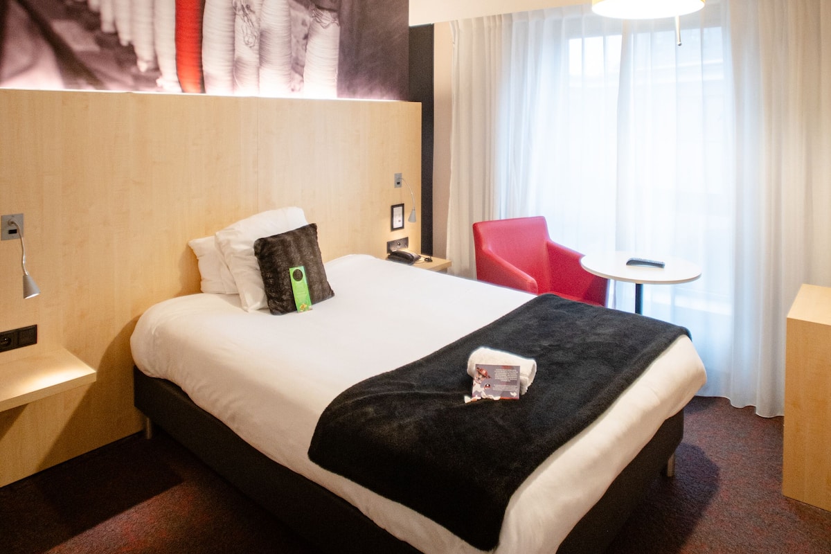 A hotel room with a large bed, a red chair, a small table, and a lamp.