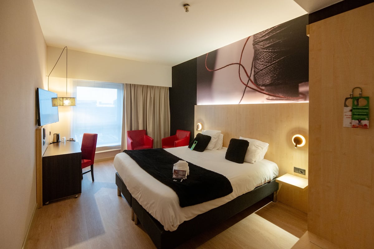 A hotel room with a large bed, a desk, a chair, a TV, and a window with curtains.