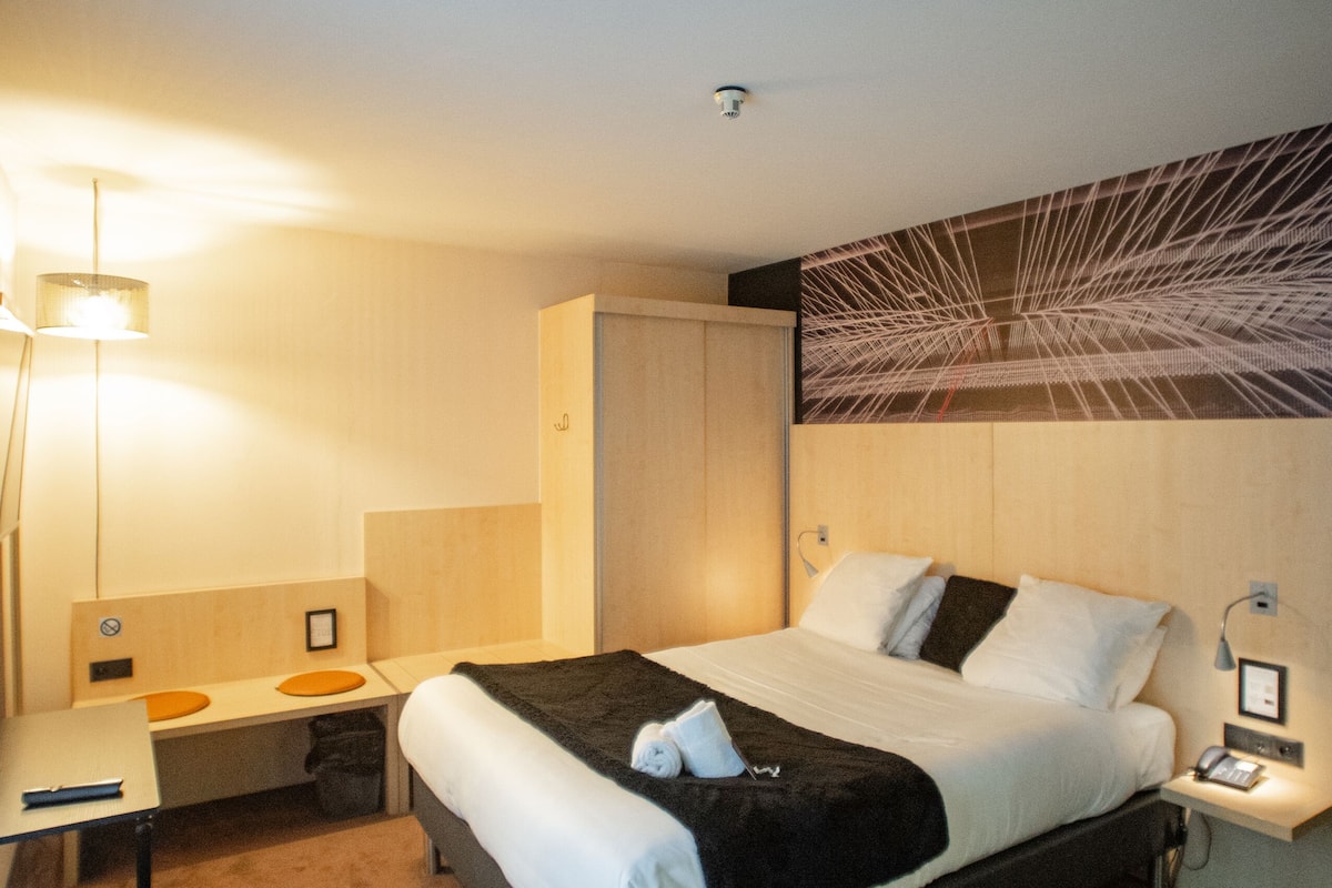 A modern hotel room with a large bed, bedside tables, a desk, and a wall-mounted artwork.