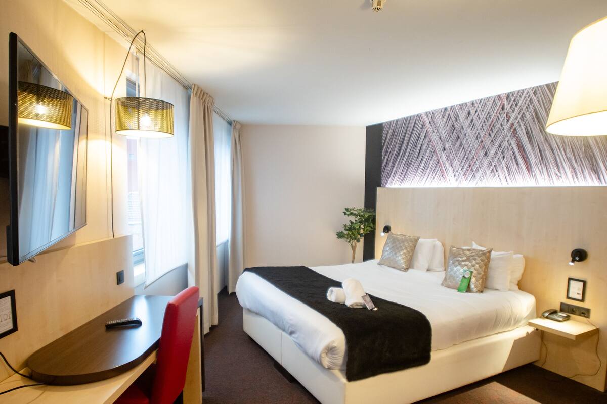 A hotel room with a bed, a desk, a chair, a lamp, and a mirror.