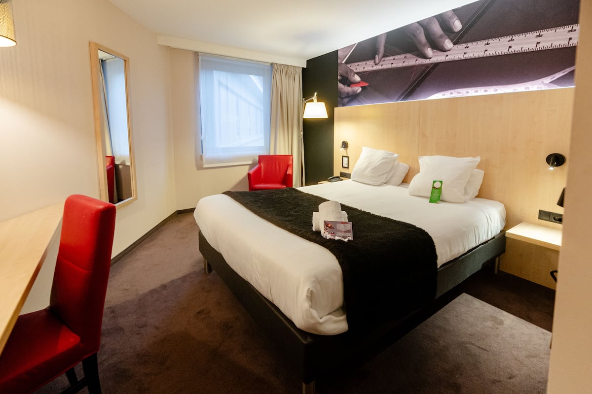 A hotel room with a bed, a red chair, a desk, a window, and a wall-mounted lamp.