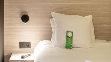A hotel room with a neatly made bed, a bedside table with a remote control, a green bottle, and a white cup on the bed.