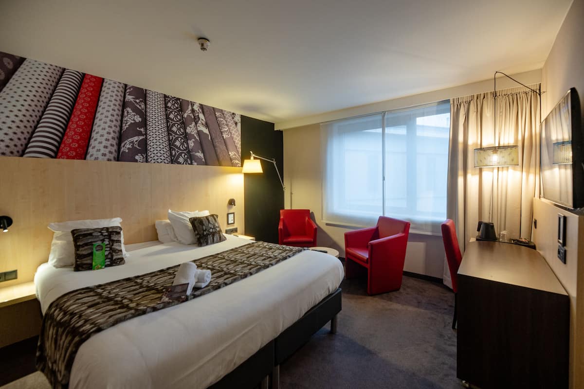 A hotel room with a bed, two red chairs, a desk, a lamp, and a large window with curtains.