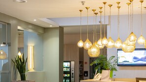 Un área de recepción moderna con un mostrador de madera, lámparas colgantes, una planta en maceta, un refrigerador con bebidas y un televisor montado en la pared.