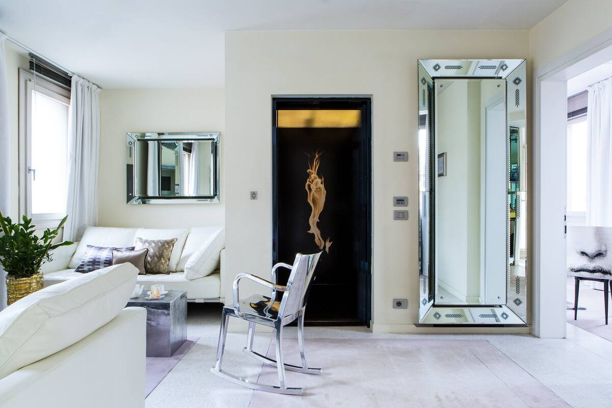 A modern living room with a white sofa, a glass coffee table, a metal rocking chair, and a large mirror.