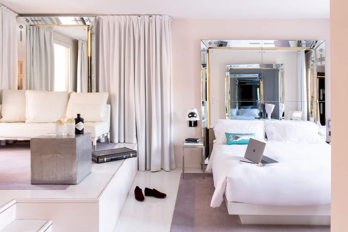 A hotel room with a bed, a sofa, a small table, a mirror, and a window with curtains.