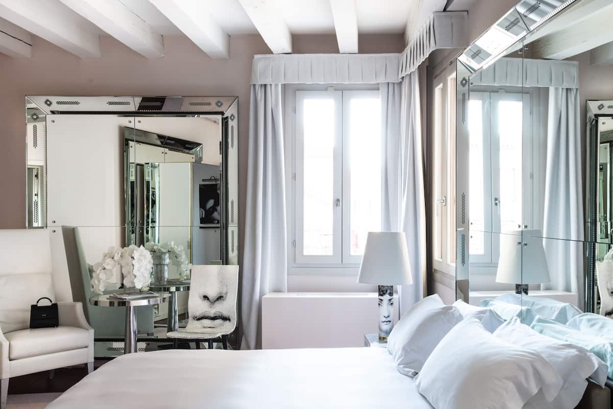 A hotel room with a large bed, a desk with a chair, a small table with a vase of flowers, and a large mirror.