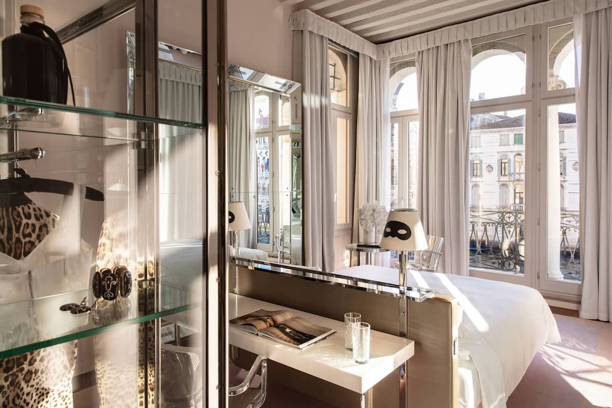 A hotel room with a glass shower enclosure, a vanity with a mirror, a bed with white linens, and a view of a cityscape through large windows.
