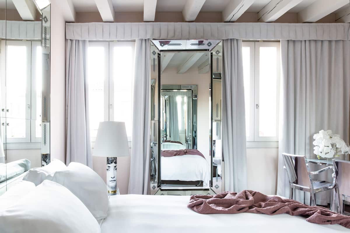 A hotel room with a bed, a desk, a chair, a lamp, and a mirror.