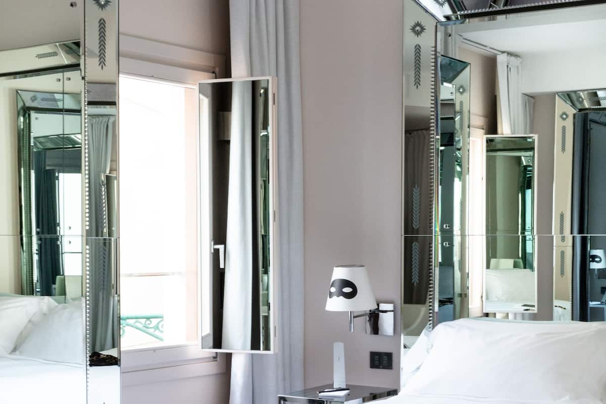 A hotel room with a large bed, a nightstand with a lamp, a mirror, and a window with curtains.