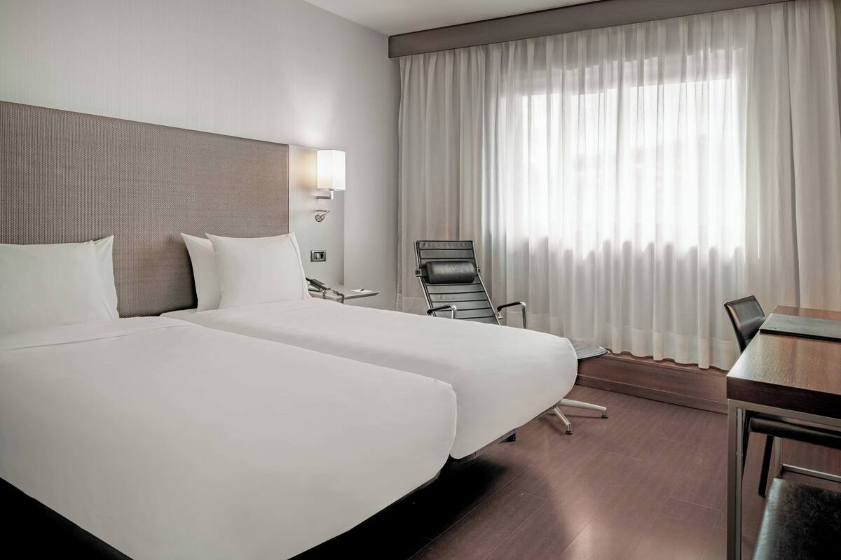 A hotel room with two beds, a desk, a chair, a lamp, and a window with curtains.