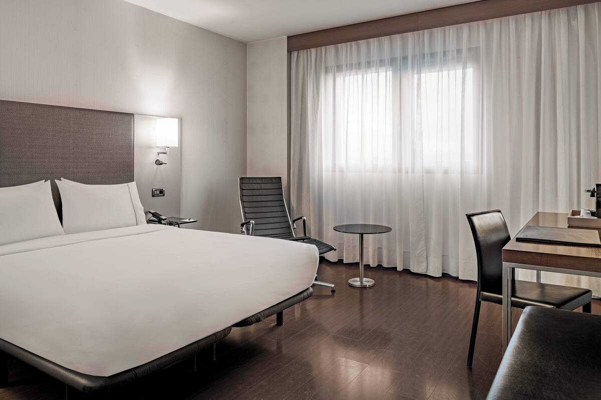 A modern hotel room with a large bed, a desk, a chair, and a small table.