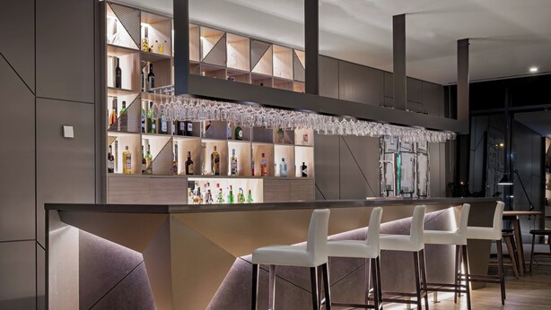 A modern bar area with a counter, bar stools, and shelves stocked with bottles. - AC Hotel Porto by Marriott (Porto)