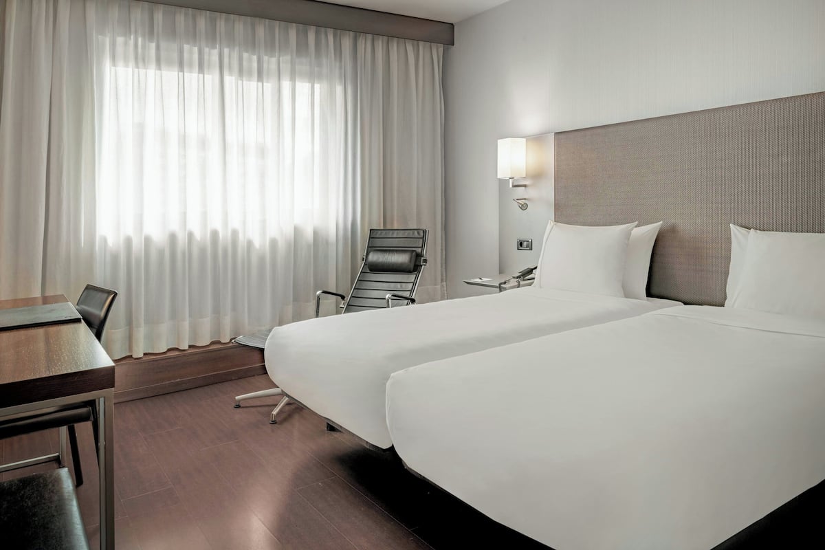 A hotel room with two beds, a desk, a chair, and a window with curtains.