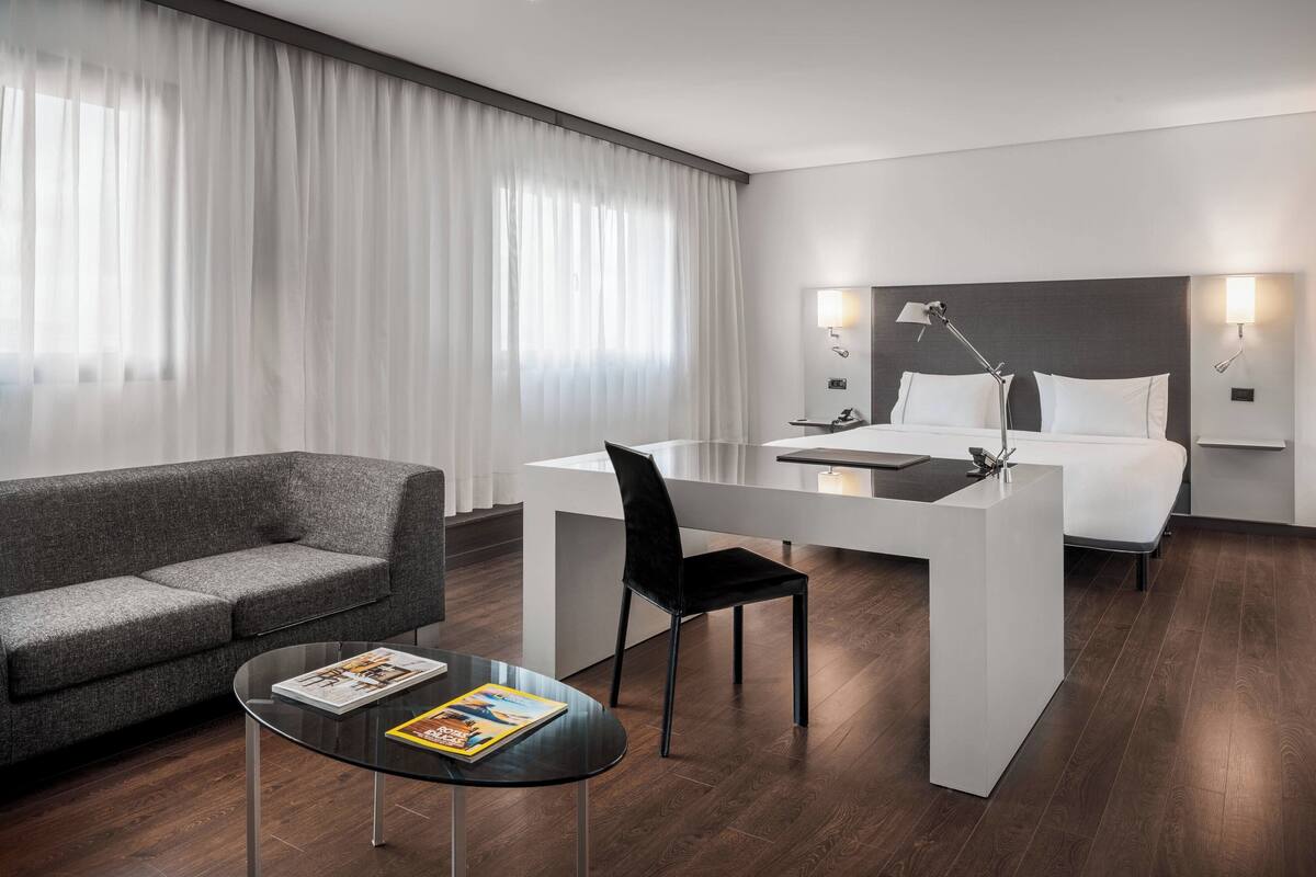 A modern hotel room with a grey sofa, a glass table with a magazine, a white desk, a black chair, a bed with white linens, and a large window with sheer curtains.