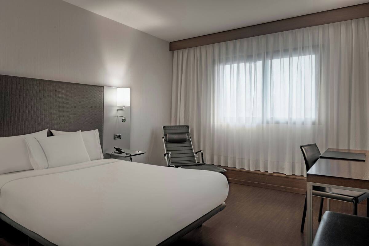 A hotel room with a large bed, a desk, a chair, a lamp, and a window with curtains.