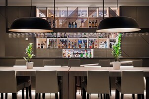 Una zona de bar con una barra alargada, varias estanterías llenas de botellas y dos grandes lámparas colgantes.