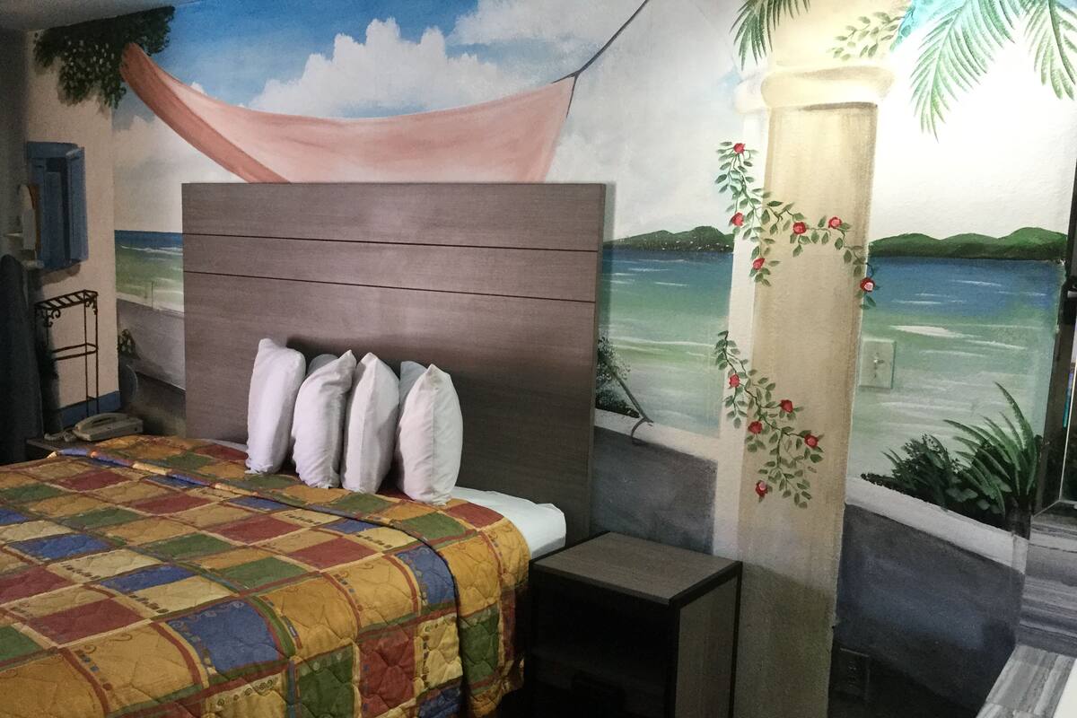 Une chambre à coucher comprenant un lit, une table de chevet, une fresque murale représentant une scène de plage et un pilier décoratif orné de fleurs.